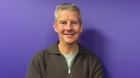 Steve Cram, a man in a grey zipped jumper is smiling at the camera against a purple background