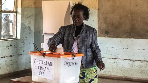 A woman in a patterned jacket and African wrap for a skirt stands at a ballot box putting her vote in. She is in an empty classroom and a blackboard can be seen behind her.