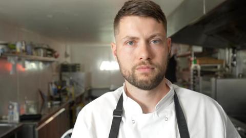 Adam Gooderham looks directly at the camera, wearing a chef's overall and black apron while standing in an industrial kitchen