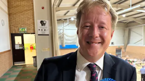 Paul Bristow smiles at the camera. He wears a dark suit jacket, a checked white shirt and a tie. He wears a Conservative party rosette. In the background is a sports centre set up for an election count.