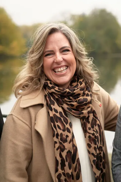 BBC/Graham Hodson Jen, who has light brown hair, is pictured standing in a park and smiling joyfully at the camera. She's wearing a tan winter coat, white top and a leopard print scarf.