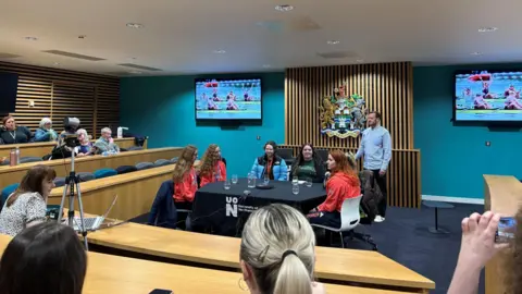 The panellists - with some wearing their red Team GB clothing - are sat at a table in a conference room, with people watching and listening. Behind them are TV screens showing rowing.