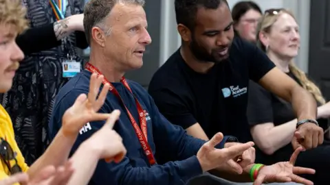 The Deaf Academy Two men are concentrating on signing the letter U in BSL with their hands. They are pointing their left index fingers to the little fingers on their right hands. Out of focus in the foreground a young man is signing the letter by pointing his right index finger to his left little finger. One is wearing a RNLI branded top and the other a branded Deaf Academy T-shirt.