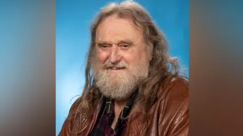 Wild Young, who ha long brown hair and a greying beard and is wearing a brown leather jacket over a dark checked shirt. The background it light blue.