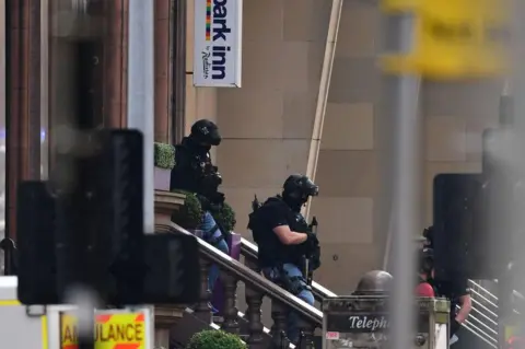 Getty Images Armed police officers in tactical gear stand on the steps outside a building near a Park Inn hotel sign, with emergency vehicles and equipment partially visible around the scene.