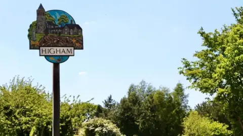 Higham's village sign. The sign features village landmarks, including the church. It is on a wooden pole. Behind it, there is blue sky, trees and shrubs.