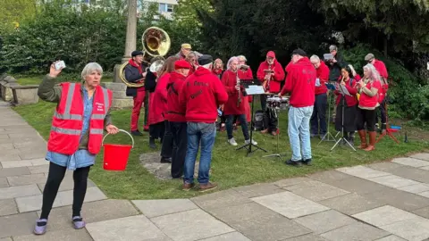 Stroud Red Band - a group of adults wearing red jumpers and hi-vis tabards - gather on a grass verge outside a church and play brass instruments.