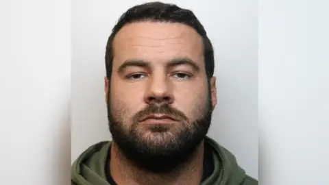 Wiltshire Police Mugshot of Oliver Cooper. He is pictured against a plain background with a blank expression. He has short dark hair and facial hair. 