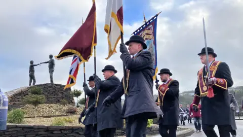 Men wearing round hats and maroon sashes are carrying a number of flags. One of which is Union Flag