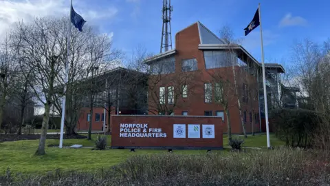 Shaun Whitmore/BBC Norfolk Police and Fire Headquarters is a large building in the middle of the image. The three storey building has several large windows, and a wall in front of the building which says its name. There are two flag poles either side of the wall.