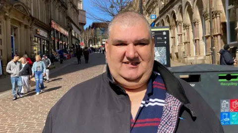 A man with a shaven head wearing a black jacket and blue polo shirt with red and white stripes standing in a street 