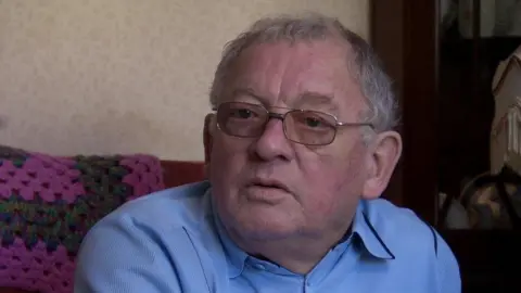 George Kirkpatrick, pictured in 2018. He has short grey hair and brown eyes, wearing a light blue shirt and glasses. He is sitting on a red sofa with a green and pink crochet blanket over it.