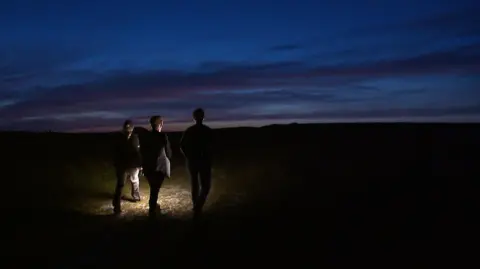 Three silhouetted figures in with their back to the camera walk across a dark landscape. The sky is dark blue and purple. The figures are holding lights which create a spotlight on the ground.