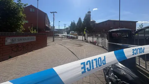 A photo of the police scene which has a blue and white police tape in the front of the picture with the word "police" written on it in blue. In the background a man, who looks like a police officer is stood in a high-vis jacket. The road name 'St Saviours Road" can be seen on a wall and below it is some grafitti.