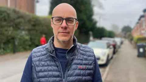 Man standing on street. He is wearing a blue jumper and sleeveless coat and glasses. Behind him are cars parked on the street which are out of focus. 