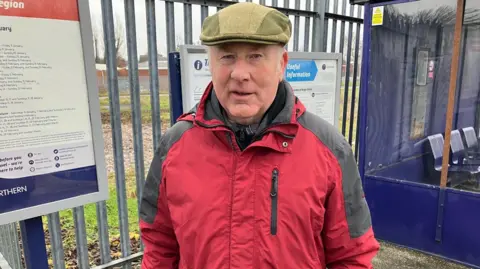 A man stands on a railway station platform in front of a grey metal fence and various noticeboards carrying timetables and other information. He has short white hair and is wearing a red jacket and a green flat cap. To the right, a waiting shelter can be seen, made of blue metal and glass sides, with blue metal seats inside.