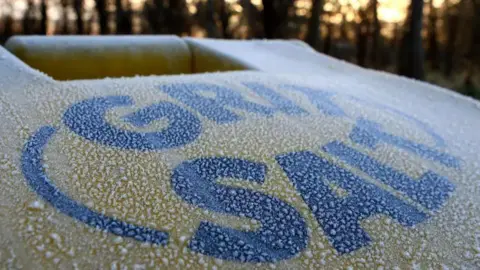 A close-up of a yellow grit salt box that is covered in a white frost. The words grit salt are in bold black writing on the top of the container. In the background there are some blurred trees and a low sun. 