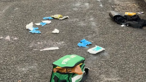 Scattered sanitary wipes, a jacket, a yellow can and a green First Aid bag on a street.