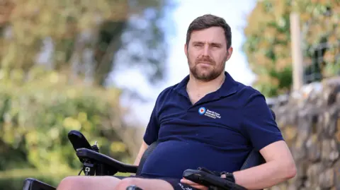 RAF Benevolent Fund/PA Real Life Rob, who is using a wheelchair and wearing a navy blue polo shirt, looks on with a neutral expression. He has short brown hair and a short beard. A stone wall and greenery is blurred in the background. 