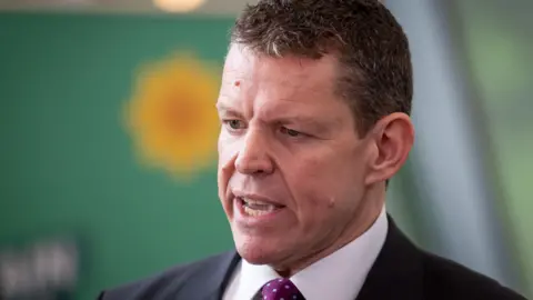 Rhun ap Iorwerth speaking off camera while being interviewed in front of a out of focus Plaid Cymru banner
