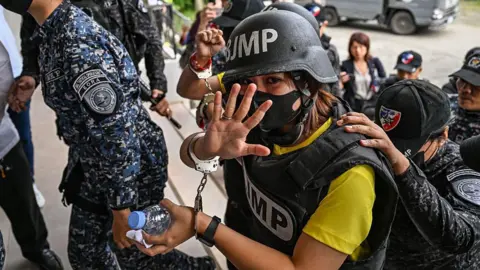A woman in a black helmet and vest, both bearing the letters BJMP, holds up five fingers of one handcuffed hand as she is escorted by figures in military uniform