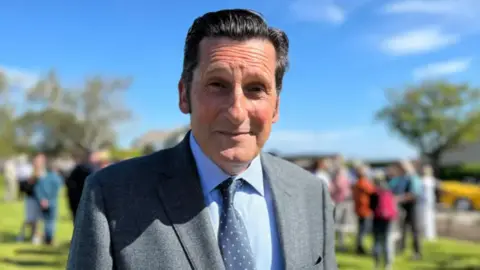 Gary Font has slicked back black hair and is wearing a grey jacket, with light blue shirt, and a navy tie with spots.