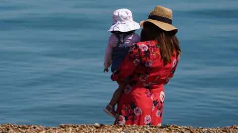 A woman is holding a young girl in her arms with their backs turned to the camera. They are on the beach looking at the clear blue water and there is shingle in the foreground. The woman is wearing a red floral dress and large floppy straw hat and the child is wearing a blue pinafore over a pink top and a white and pink patterned sun hat.