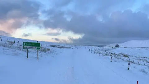 A road on the mountain which is covered in snow. There is a green sign that points the direction to both Douglas and Kirk Michael