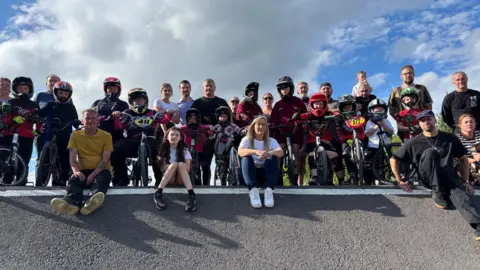 A large group of BMX riders holding their bikes and wearing helmets stand at the top of a ramp alongside other adults.