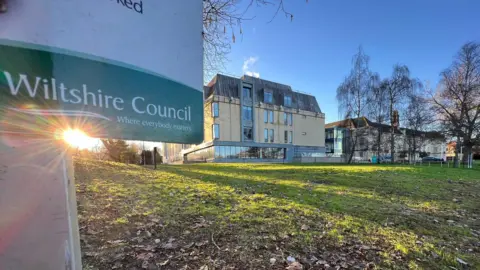 Bottom of a Wiltshire Council sign showing a logo with sun shining behind. You can see the headquarters building in the background across a lawn.
