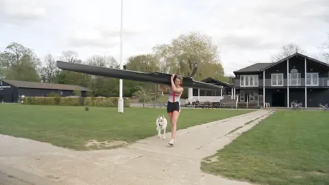 Pippa carrying her rowing boat on her shoulder away from her rowing clubhouse. She is being closely followed by her white rescue dog.