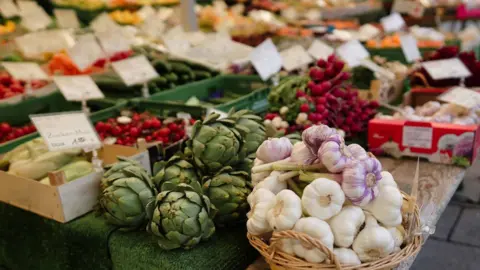 Getty Images A fruit and veg stall on a market with a basket of garlic heads in the foreground as well as artichokes. In the background and out of focus you can see rows of other fresh produce including radishes and tomatoes.