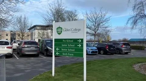 Ladies' College Guernsey. A sign in the foreground with cars parked behind and then buildings behind that.