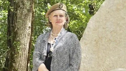 Joanna Burri-Weaver Joanna Burri-Weaver wears a military beret and stands next to a stone memorial in a forest