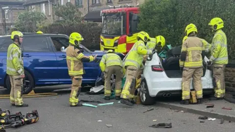 A group of firefighters surround a white car on a residential street, which has been heavily damaged. In the background, a fire engine can be seen.