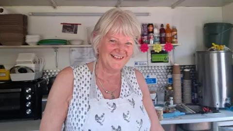 'It's a dying town': Community hit hard by factory's loss 9 BBC/Seb Cheer A woman with white hair looks into the camera, smiling. She is wearing a light blouse and an apron, and is standing in a sandwich van. Behind her are an oven, disposable containers and cups, and a number of cold drinks.