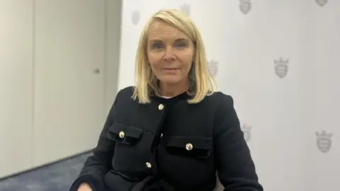 A woman with should length blonde hair and blue eyes looking directly at the camera. Her face is dead-panned. She is wearing a black coat with gold buttons. Behind her is a white wall with grey States of Jersey logos on it. The carpet is blue. 