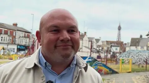 An image of councillor Dave Flanagan standing next to a playground in Revoe.  He is wearing a blue shirt and cream jacket, in the distance you can see Blackpool tower.