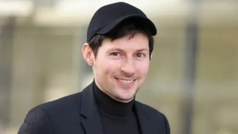 Pavel Durov, a white man white short black hair. He has designer stubble. He is wearing all black, including a cap, a turtleneck and a suit jacket.