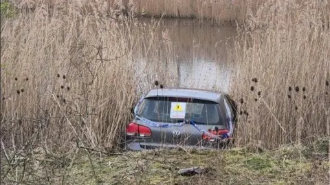 Leicestershire Fire and Rescue Service A VW Golf in some water with reeds either side of the car. There is police tape on the back of the car.