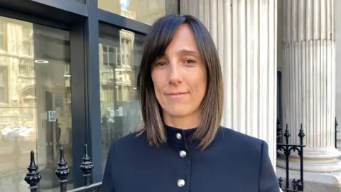 Debbie Hatch standing outside of Bristol Crown Court. She is wearing a navy blue jacket with round silver buttons. She has short brown straight hair.