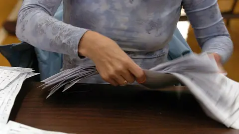 A person handles a sheaf of ballot papers while seated at a desk.