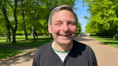 Paul is smiling widely into the camera He has short greying hair and is wearing a white shirt under a black cardigan. He's standing in a park on a bright sunny day. 