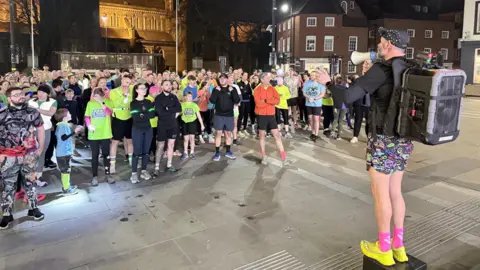 A group of people in bright neon clothing stand in a crowd and a man at the front is in colourful shorts, bblack top, a cap and have a speaker on his back