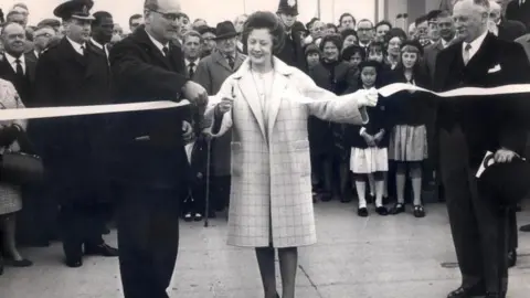 PA Media A black and white photo shows a woman in a large overcoat cutting a ribbon, with two gentlemen in smart suits and a bowler hat, with a crowd of onlookers of all ages behind them in smart dress and some in uniforms