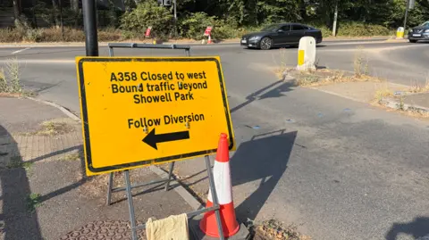 A yellow sign is pictured by a road diverting traffic due to a road closure. There is a cone next to the sign and two cars can be seen using the road in the background.