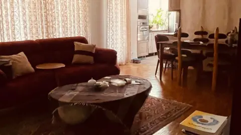 Social media Living room with a sofa, coffee table and rug, with the curtains closed, and a dining table and kitchen in the background.