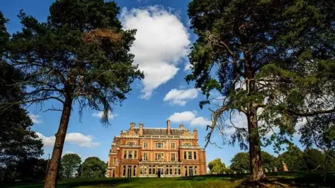 A distant shot of the outside of Rossington Hall's building, with a green door and reddish brick buildings, and trees surrounding it