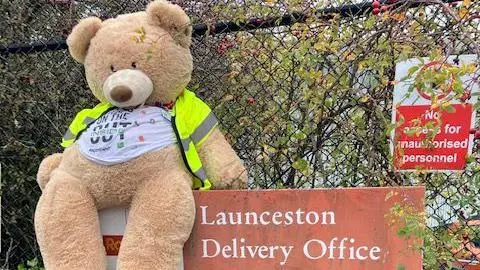 Nikki Gaynor A very large cuddly beige-furred bear is wearing a yellow high visibility vest and is sitting on the sign at the Royal Mail's Launceston Delivery Office. Behind it is a wire fence with hedge plants weaving through it. On the fence is a slightly overgrown red and white sign reading: "No access for unauthorised personnel."