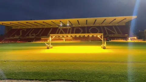 Green astroturf with an illuminated golden circle from floodlights. Stands are in the background with "SWINDON" written across the chairs.